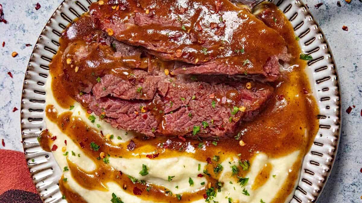 A plate with slices of braised beef covered in brown gravy, served alongside creamy mashed potatoes, garnished with chopped parsley and red pepper flakes.