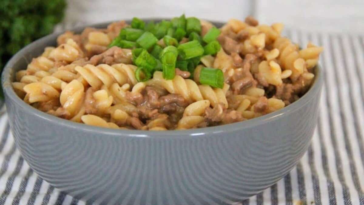 A gray bowl filled with creamy rotini pasta and ground beef, topped with chopped green onions, sits on a striped cloth.