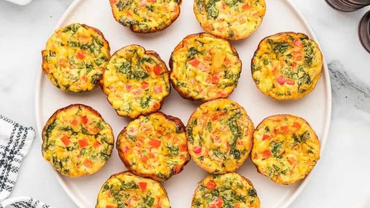 A plate of twelve colorful egg muffins with visible pieces of spinach, red peppers, and other vegetables, arranged neatly in a circle on a white surface.