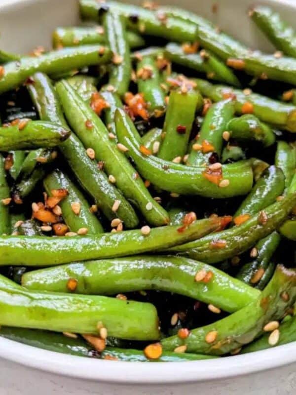 A white bowl filled with sautéed green beans garnished with sesame seeds and spices, sitting on a light-colored surface.