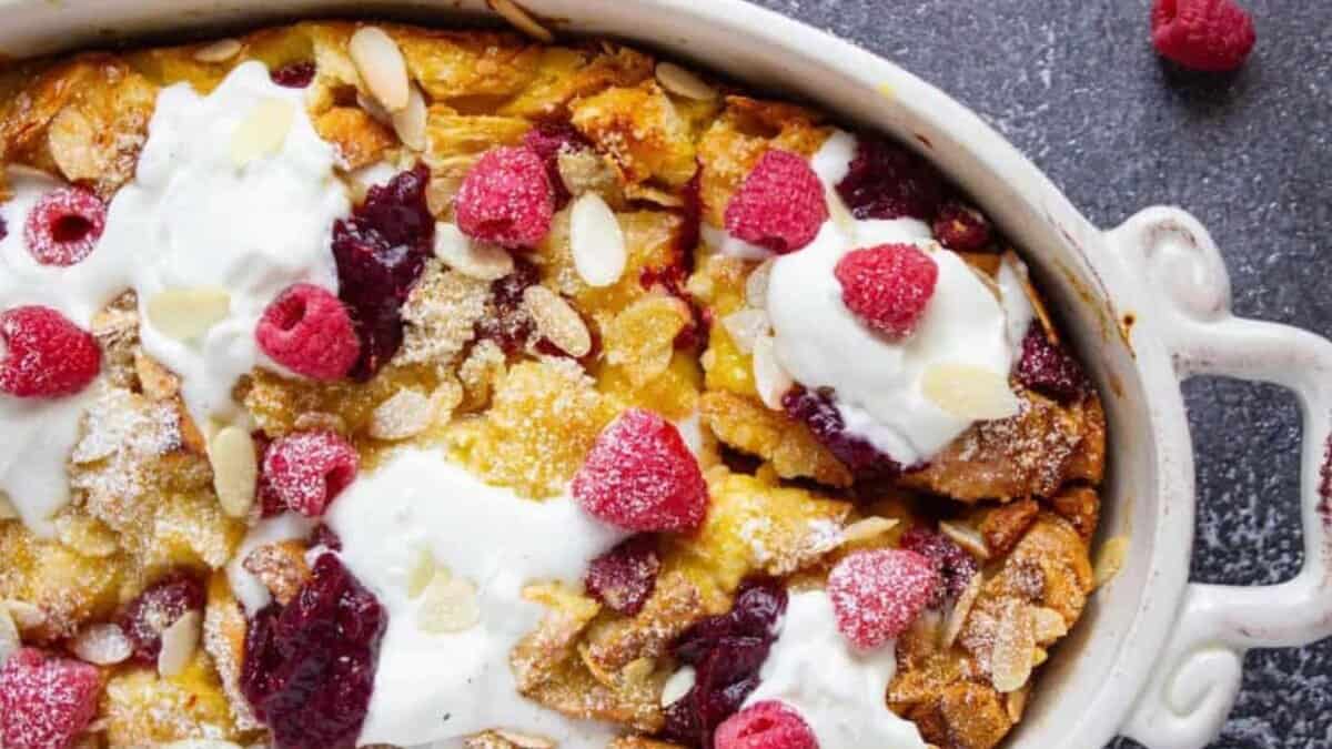 A baked bread pudding topped with whipped cream, raspberry sauce, fresh raspberries, sliced almonds, and powdered sugar in a white oval dish on a dark surface.