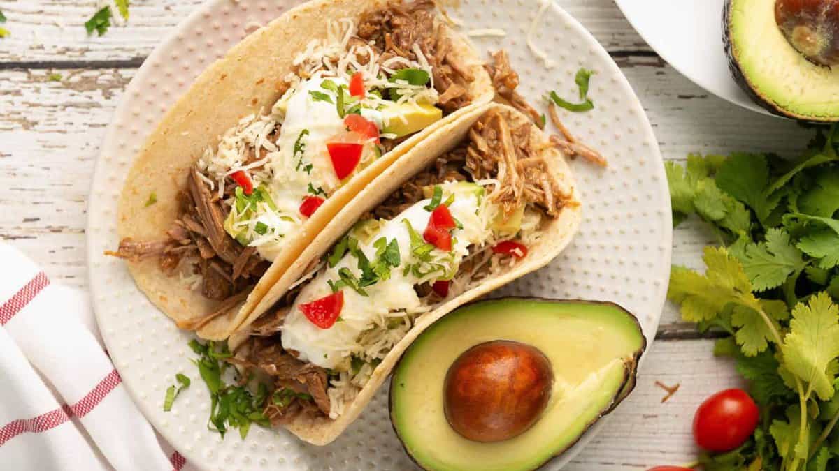 Two shredded beef tacos topped with sour cream, shredded cheese, chopped tomatoes, and cilantro on a white plate next to half an avocado. Fresh cilantro, avocado, and tomatoes are in the background.