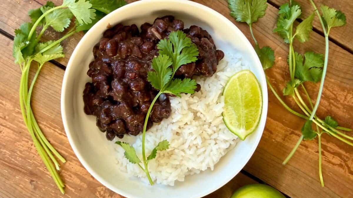 A white bowl filled with black beans and white rice, garnished with cilantro and a lime wedge, sits on a wooden surface surrounded by fresh cilantro and a whole lime.
