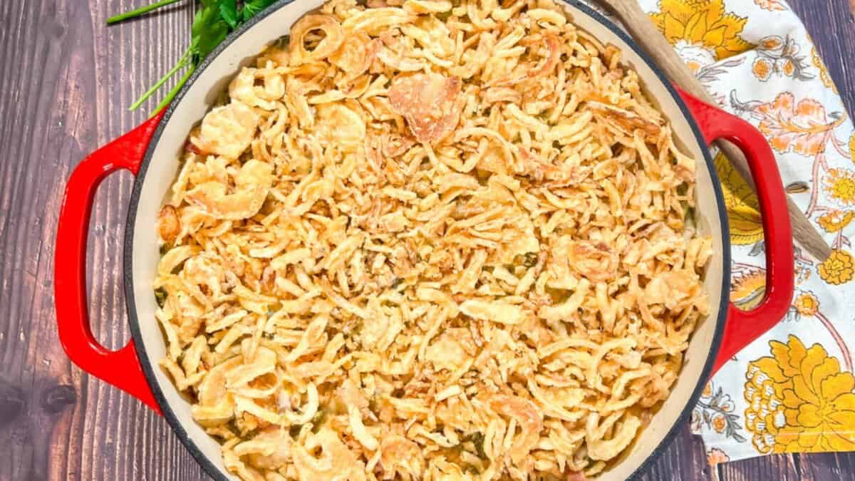 A red casserole dish filled with green bean casserole topped with crispy fried onions sits on a wooden table next to a floral napkin and a green leafy garnish.