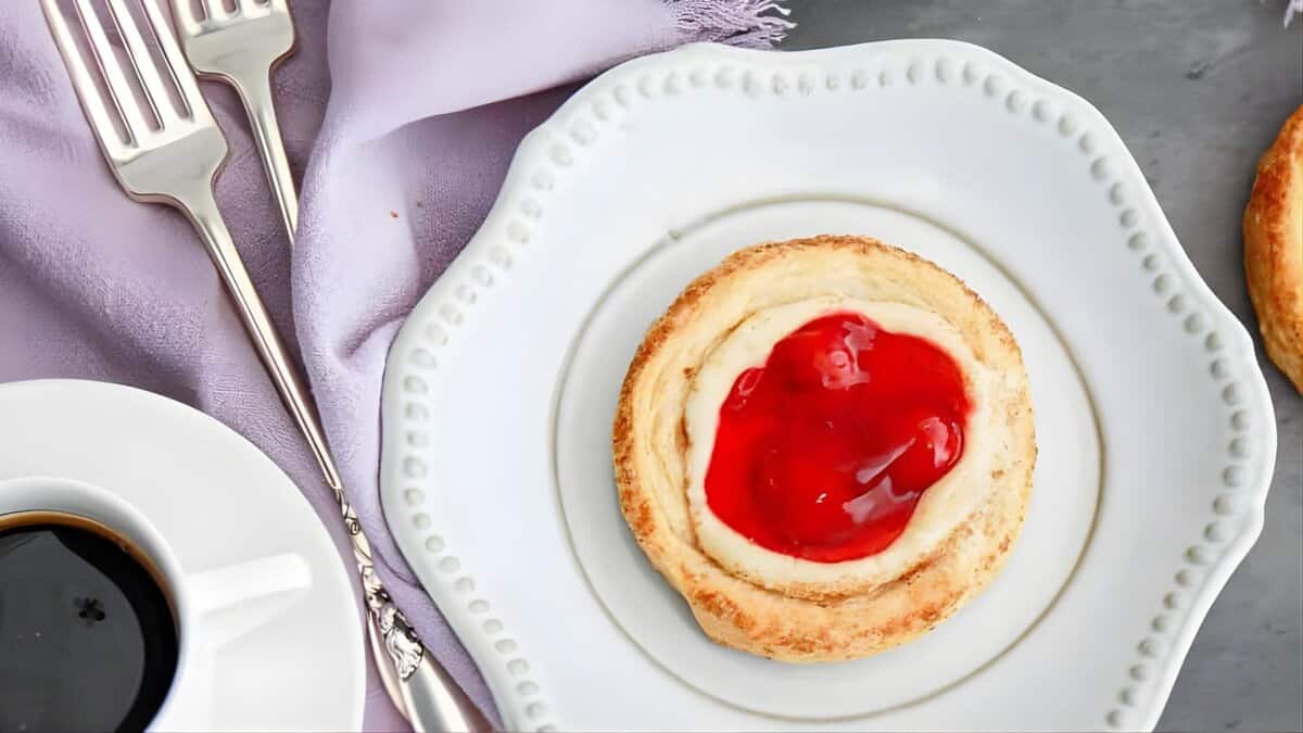 A pastry topped with bright red jelly sits on a white scalloped plate beside a cup of coffee, two forks, and a lavender napkin on a gray surface.
