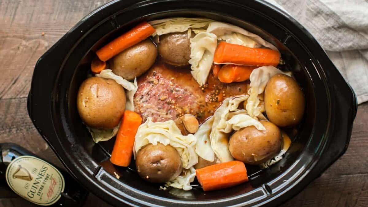 A slow cooker filled with whole red potatoes, carrot pieces, cabbage wedges, and a seasoned meat brisket in broth. A bottle labeled "Guinness" is partially visible beside the cooker on a wooden surface.