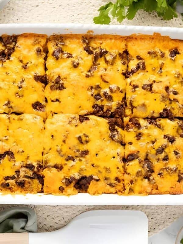 A baked casserole topped with melted cheese and ground beef, cut into squares in a white dish. Surrounding the dish are bowls of shredded lettuce, diced tomatoes, cilantro, sour cream, and plates with forks.