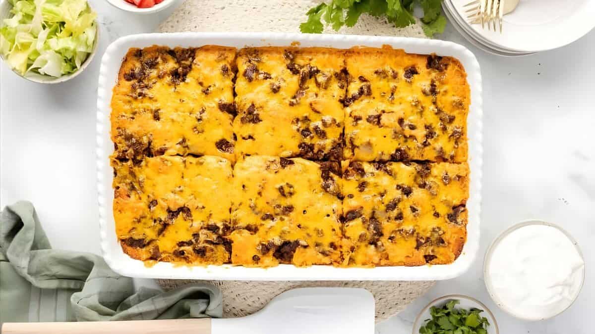 A baked casserole topped with melted cheese and ground beef, cut into squares in a white dish. Surrounding the dish are bowls of shredded lettuce, diced tomatoes, cilantro, sour cream, and plates with forks.