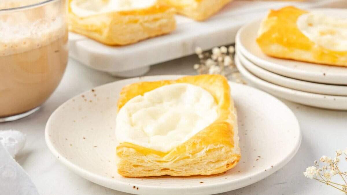 A square cream cheese Danish pastry sits on a white plate, with a cup of coffee and more pastries in the background on a white surface. The pastry has golden, flaky layers and a creamy white filling in the center.
