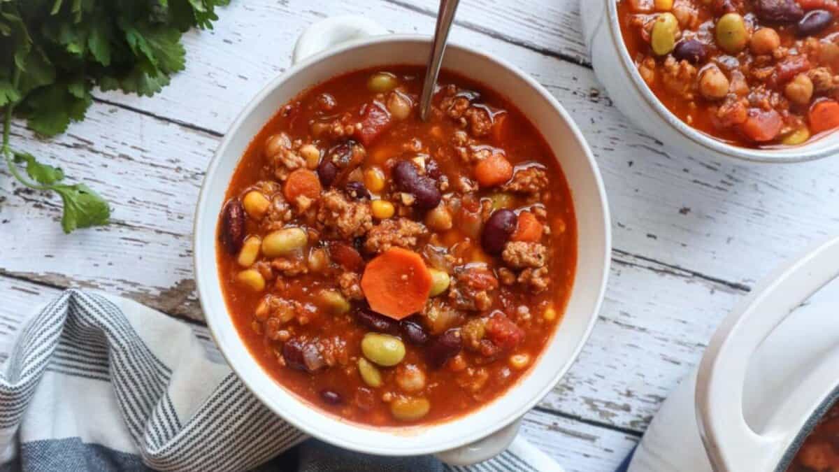 A bowl of hearty vegetable and bean chili, filled with corn, carrots, kidney beans, and ground meat, sits on a rustic wooden table with a spoon and a striped napkin beside it. Another bowl is partially visible.