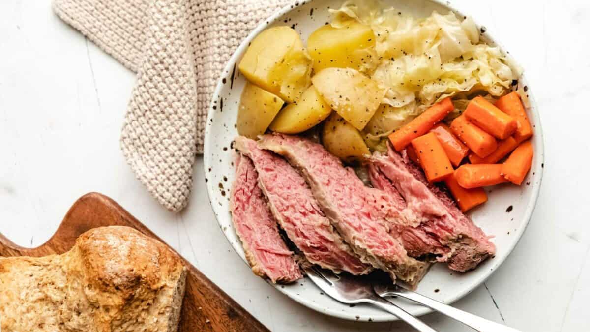 A plate with slices of corned beef, boiled potatoes, cabbage, and carrots sits on a white surface, next to a piece of bread and a fork and knife.