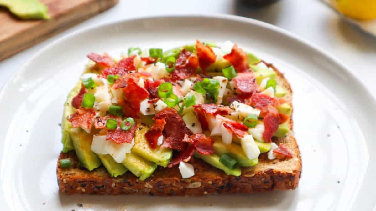 A slice of multigrain toast topped with sliced avocado, chopped boiled egg, crispy bacon pieces, and sliced green onions, served on a white plate.