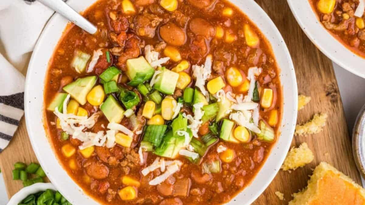 A bowl of chili topped with chopped green onions, shredded cheese, and corn, with cornbread and more green onions on the side. The chili contains beans, corn, ground meat, and tomatoes.