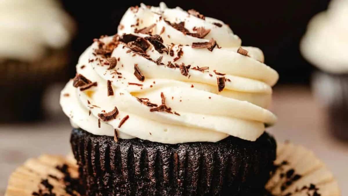 A close-up of a chocolate cupcake with creamy white frosting, topped with chocolate shavings, in an opened brown paper liner.