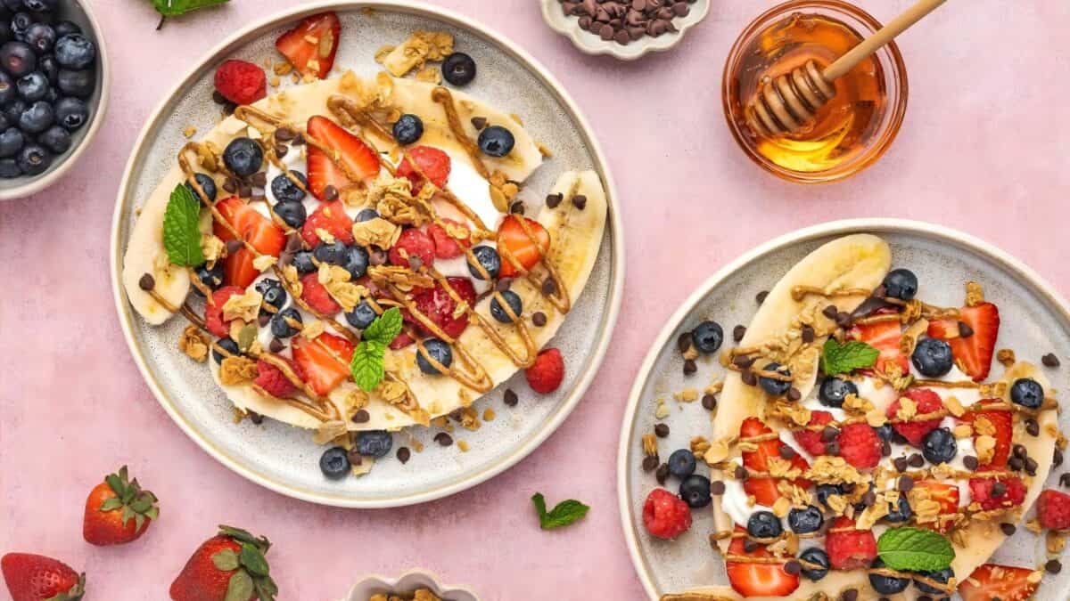 Two plates with banana splits topped with strawberries, blueberries, granola, chocolate chips, and a drizzle of honey, surrounded by bowls of blueberries, strawberries, chocolate chips, and a jar of honey on a pink surface.