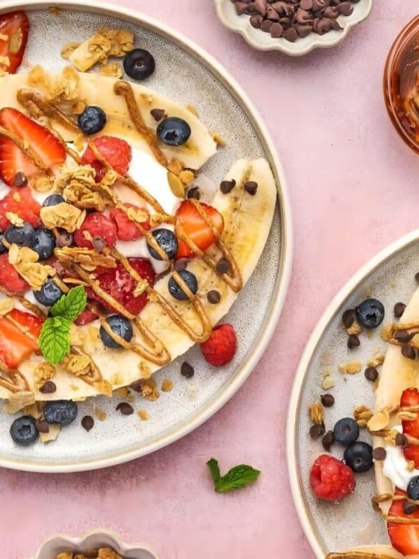 Two plates with banana splits topped with strawberries, blueberries, granola, chocolate chips, and a drizzle of honey, surrounded by bowls of blueberries, strawberries, chocolate chips, and a jar of honey on a pink surface.