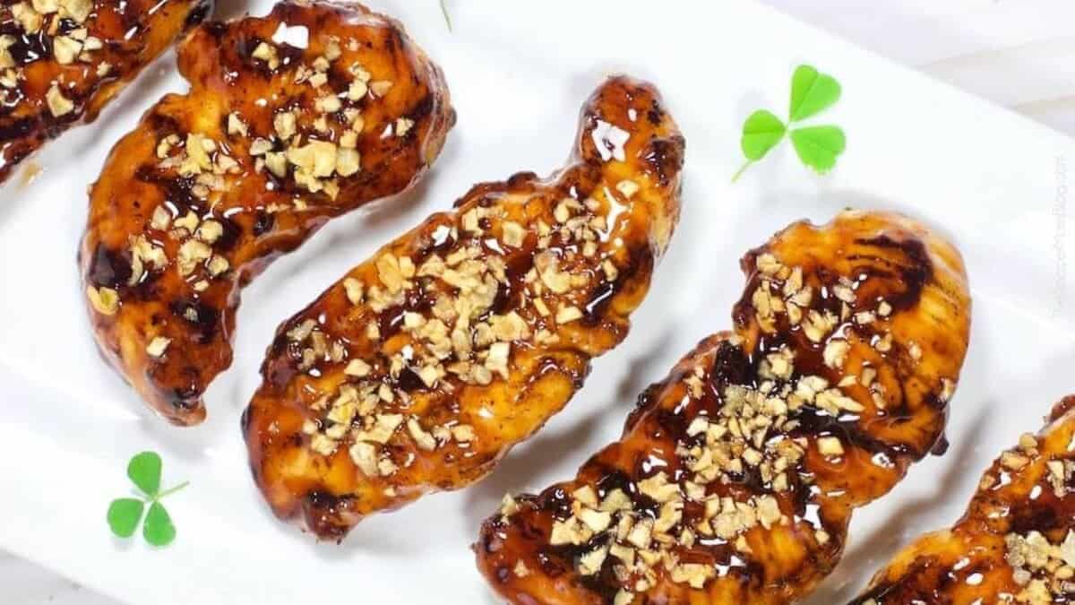 Five pieces of glazed grilled chicken topped with chopped nuts are arranged on a white rectangular plate, garnished with a few small green clover leaves.