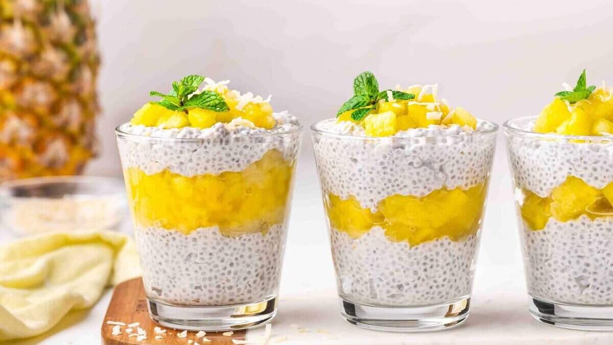 Three glasses of chia pudding layered with bright yellow mango puree and topped with fresh mint leaves, set on a white surface with a blurry pineapple and ingredients in the background.