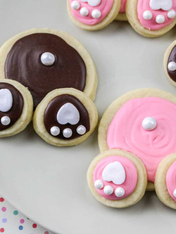 Festive cookies decorated with pink and brown icing to resemble bunny paws, placed on a white plate with a colorful polka dot background. Each paw features heart and pearl-like details, making them perfect Easter cookies.