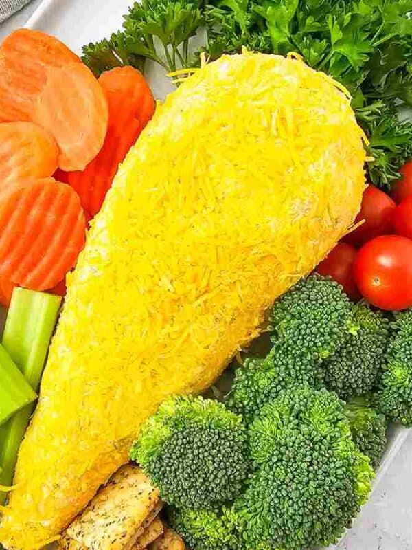A platter with sliced carrots, celery sticks, broccoli, cherry tomatoes, crackers, leafy greens, and a chicken drumstick-shaped cheese ball coated in shredded cheese—perfect for festive Easter side dishes.