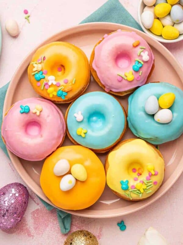 A plate of colorful frosted donuts in pink, yellow, and blue, decorated with sprinkles and mini egg candies. Surrounded by Easter eggs and additional donuts on pastel surfaces, these festive desserts brighten any celebration.