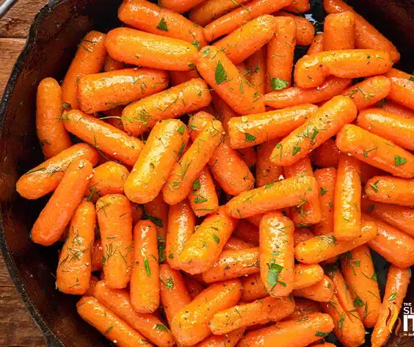 A skillet filled with glazed baby carrots garnished with chopped herbs, surrounded by a spoon and a bowl of honey on a wooden table—perfect for Easter side dishes. The words “The Slow Roasted Italian” appear in the bottom right corner.