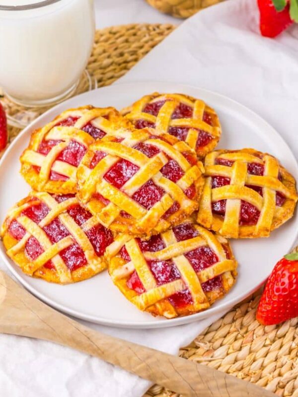 A plate of mini lattice strawberry pies sits on a woven placemat, surrounded by fresh strawberries, a wooden spoon, and a glass of milk. The pies have a golden, crisscrossed crust and a bright red filling.
