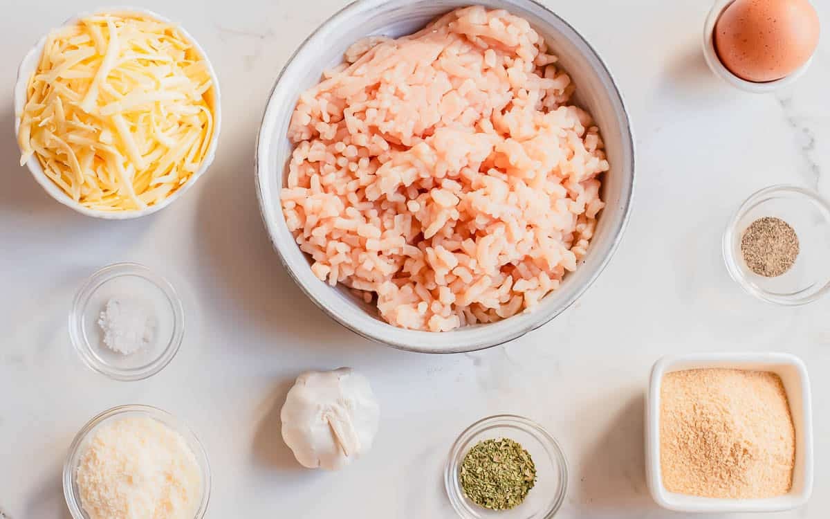 A bowl of ground chicken is surrounded by shredded cheese, an egg, salt, pepper, grated cheese, garlic, dried herbs, and breadcrumbs on a white surface.