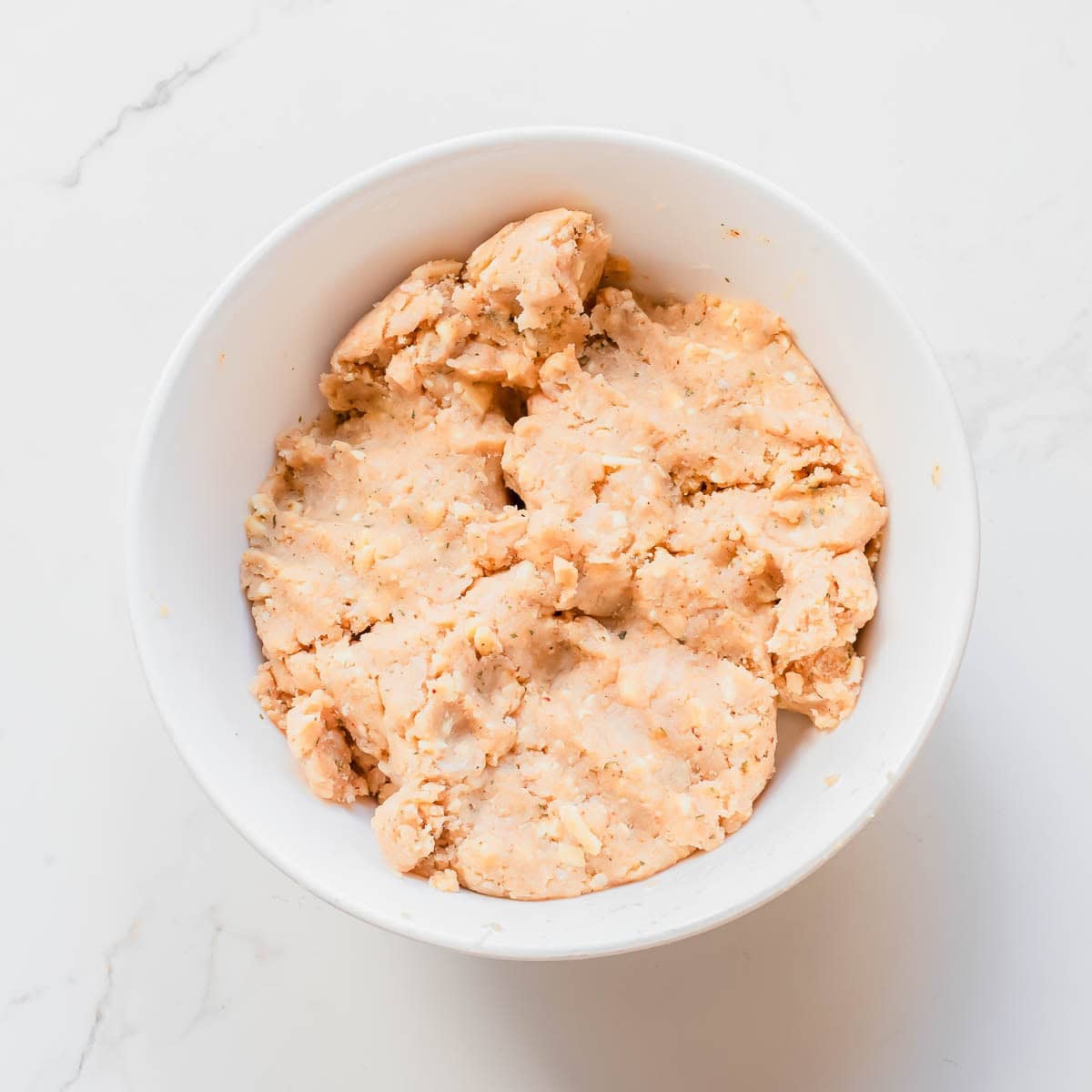 A white bowl filled with a ground chicken meatball mixture sits on a white marble surface.