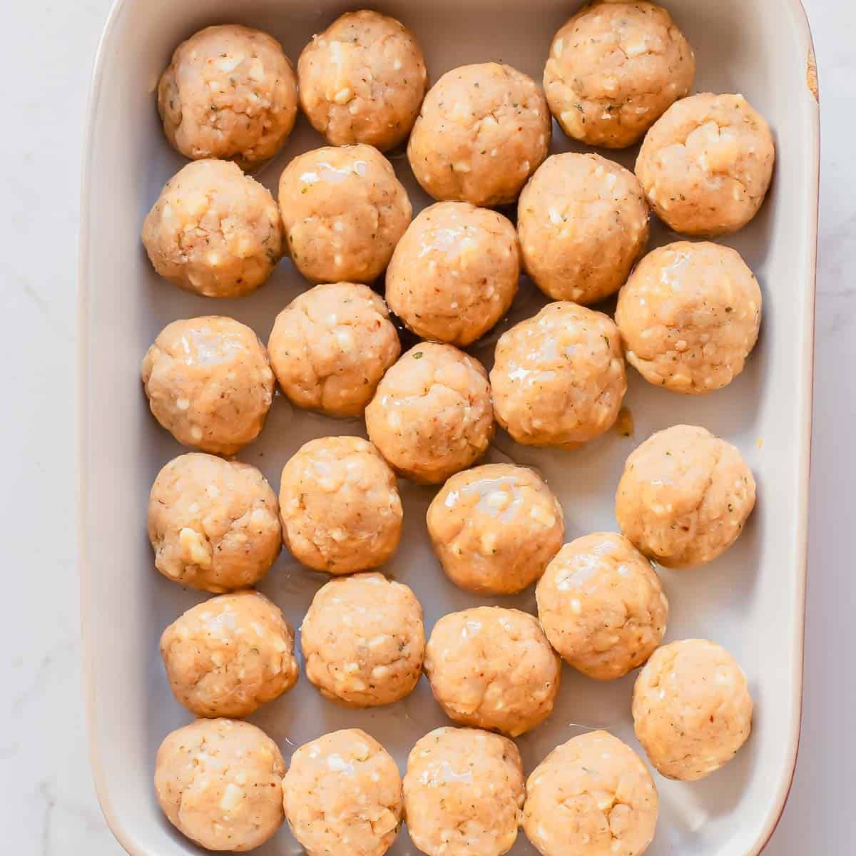 A white baking dish filled with evenly spaced, uncooked chicken meatballs on a white marble surface.