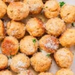 A close-up view of several baked chicken meatballs sprinkled with grated parmesan cheese and garnished with chopped parsley on a white surface.