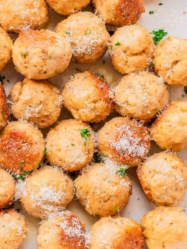 A close-up view of several baked chicken meatballs sprinkled with grated parmesan cheese and garnished with chopped parsley on a white surface.
