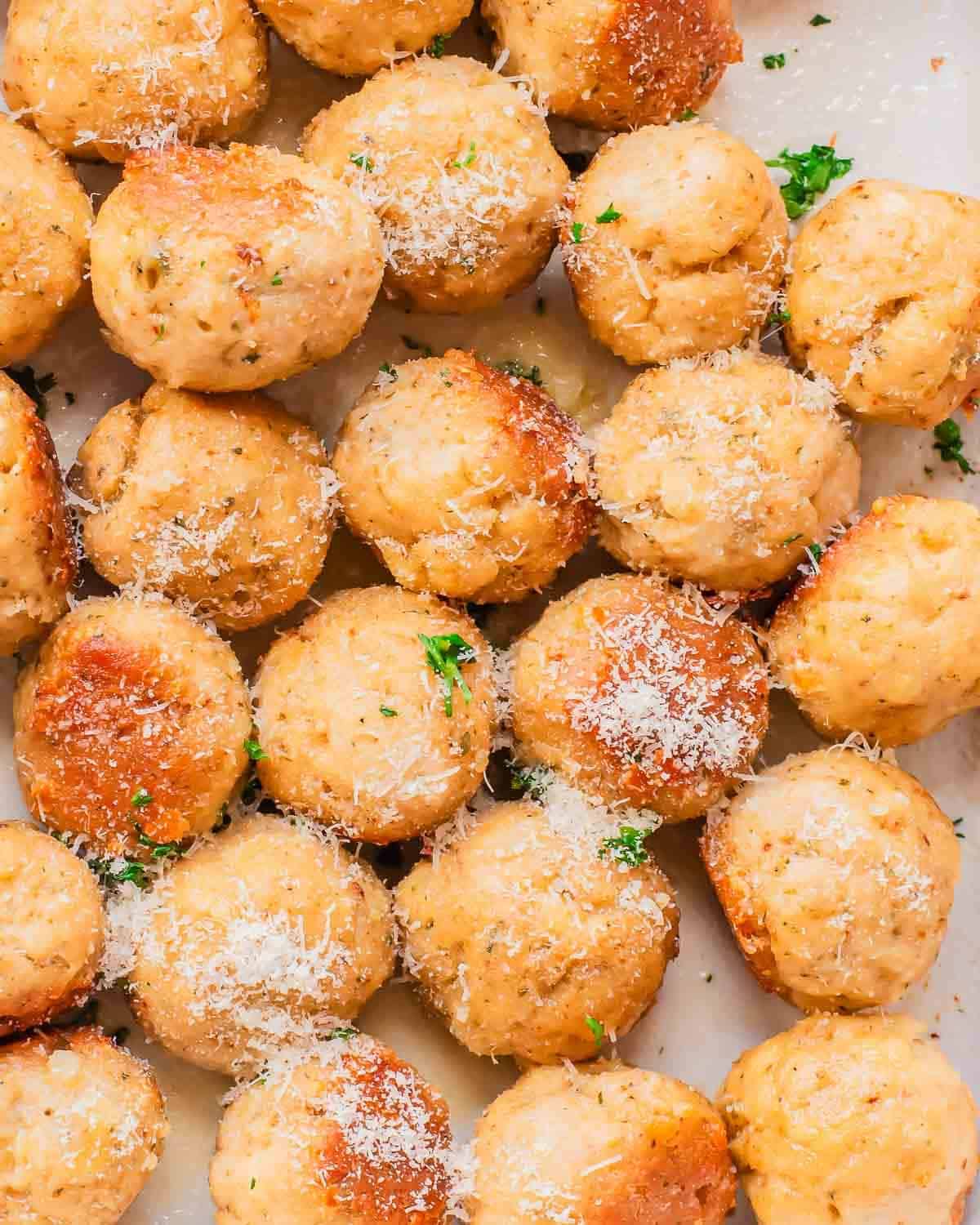 A close-up view of several baked chicken meatballs sprinkled with grated parmesan cheese and garnished with chopped parsley on a white surface.