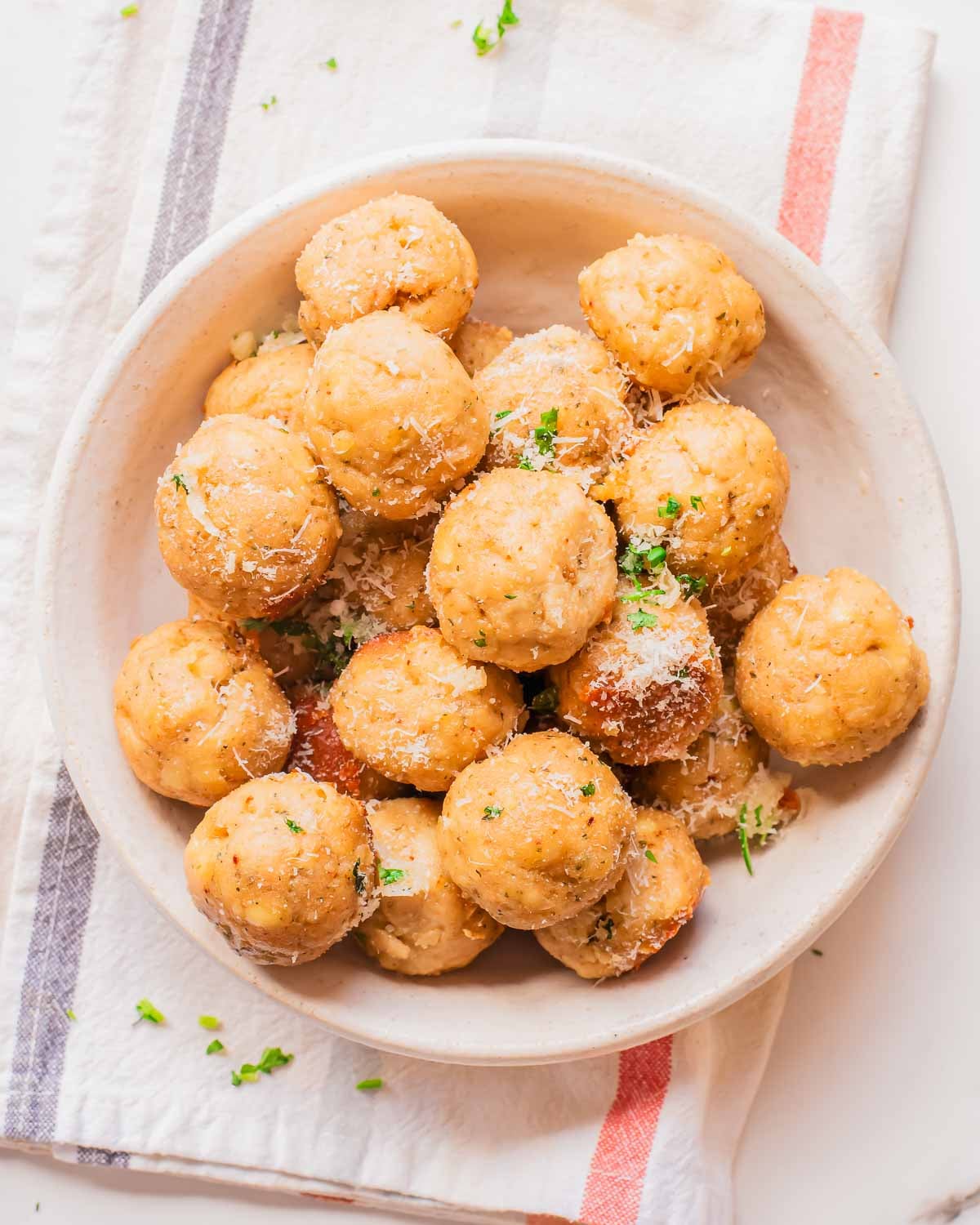 A bowl filled with round, golden-brown cheesy chicken meatballs, garnished with grated cheese and chopped parsley, sits on a plate over a striped cloth napkin.