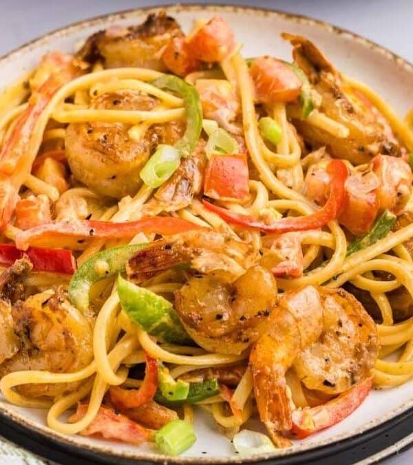 A plate of creamy pasta topped with shrimp, sliced red bell peppers, green onions, and spices—a delicious example of comfort food—served with a fork and knife beside it on a white table.