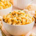 A white bowl filled with creamy, cheesy macaroni and cheese with cauliflower, garnished with chopped herbs, sits on a light-colored surface with scattered pasta and cauliflower pieces in the background.