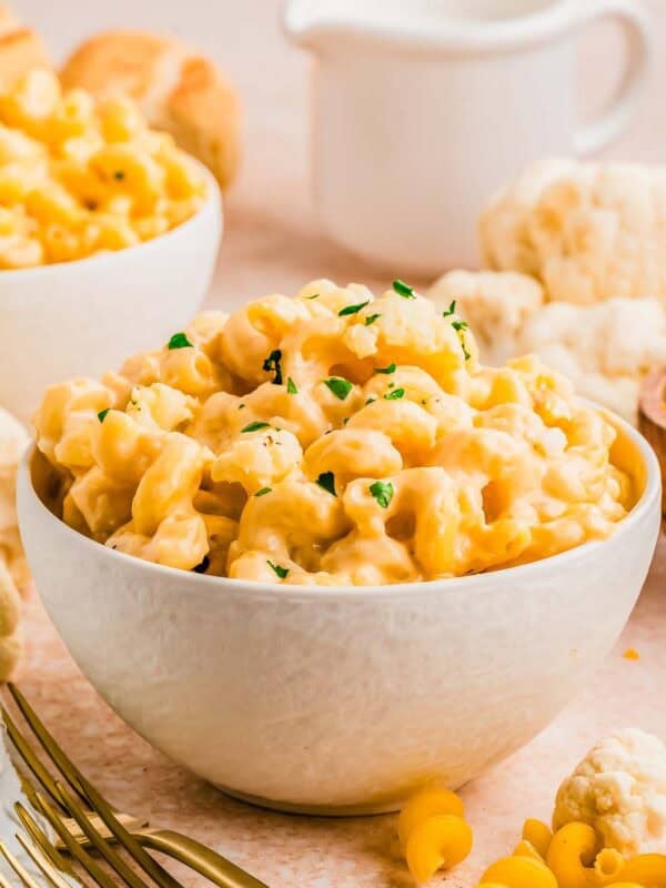 A white bowl filled with creamy, cheesy macaroni and cheese with cauliflower, garnished with chopped herbs, sits on a light-colored surface with scattered pasta and cauliflower pieces in the background.
