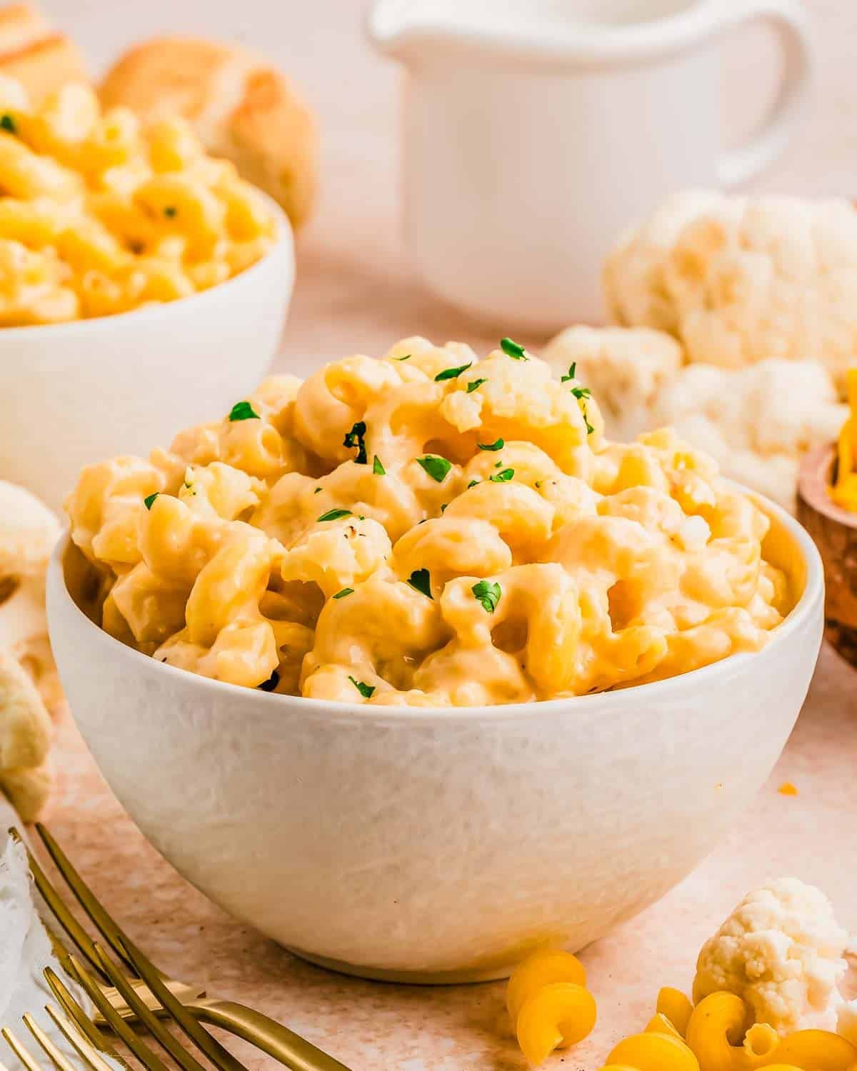 A white bowl filled with creamy, cheesy macaroni and cheese with cauliflower, garnished with chopped herbs, sits on a light-colored surface with scattered pasta and cauliflower pieces in the background.