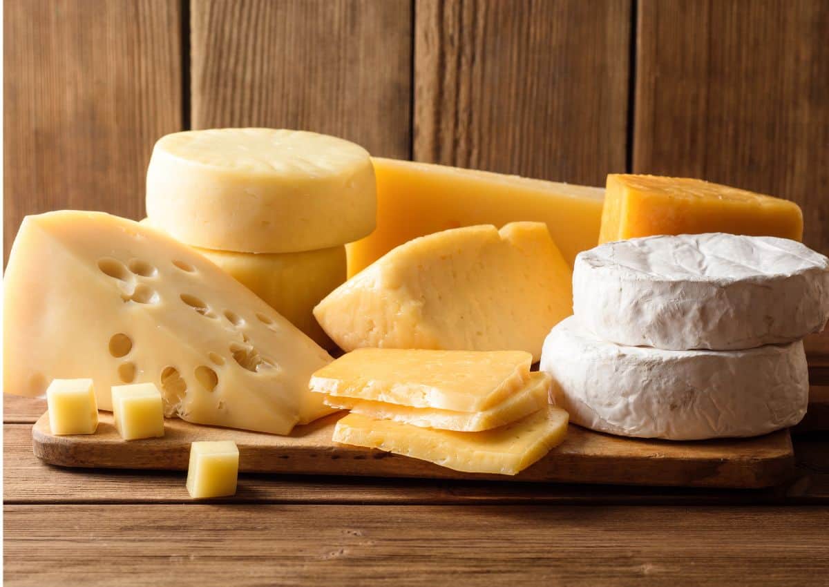 A wooden board with various cheeses&mdash;wheels, wedges, slices, blocks, and cubes&mdash;arranged in front of a rustic wooden background. Perfect for your next Aldi shopping haul or to feature in your latest Aldi review. Some cheeses have holes and some have a white rind.