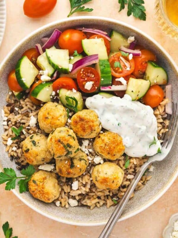 A bowl filled with brown rice, chicken meatballs, tomato-cucumber salad, feta cheese, and tzatziki sauce, garnished with parsley—perfect for easy recipes or chicken dinners. Nearby are more meatballs, cherry tomatoes, tzatziki, and drinks.
