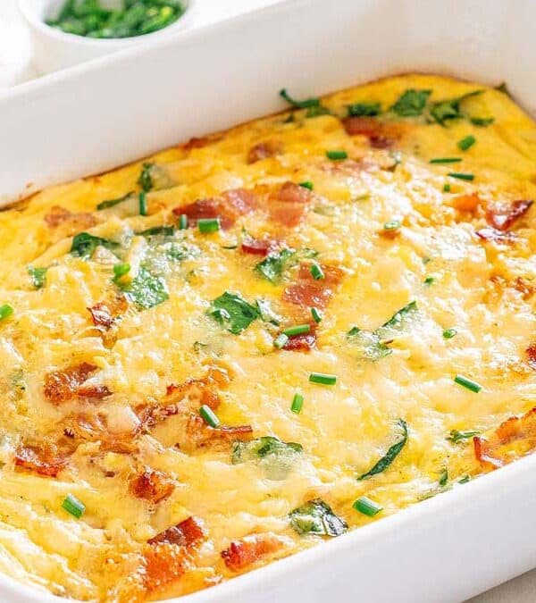 Baked egg casserole in a white dish, topped with chopped green herbs, melted cheese, visible spinach, and pieces of cooked bacon—a perfect easy breakfast for stress free mornings. Nearby are ingredients like cheese, eggs, and fresh greens.