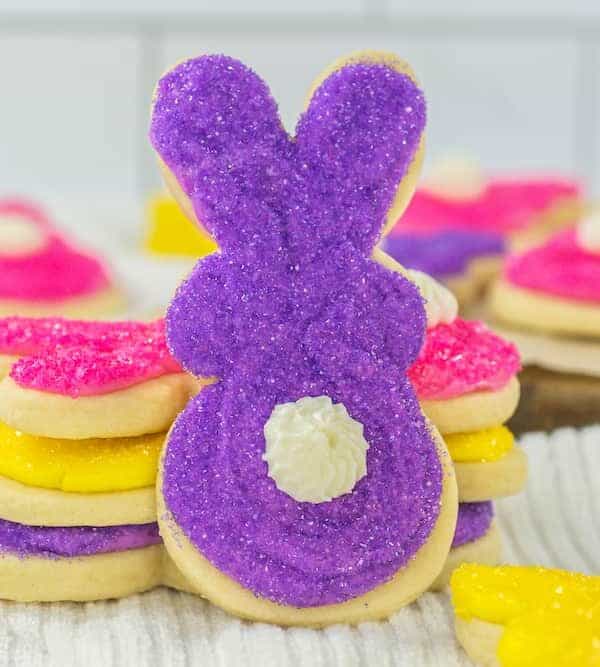 A bunny-shaped sugar cookie perfect for spring celebrations is decorated with purple sugar and a white icing tail, stacked atop pink and yellow bunny cookies, all displayed on a white cloth with a blurred kitchen background.