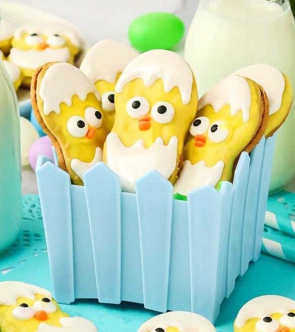 Adorable Cookies shaped like baby chicks hatching from eggs are arranged in a blue picket fence container. Bottles of milk and pastel Easter eggs add a festive touch, with a blue cloth and straws completing this charming Easter scene.