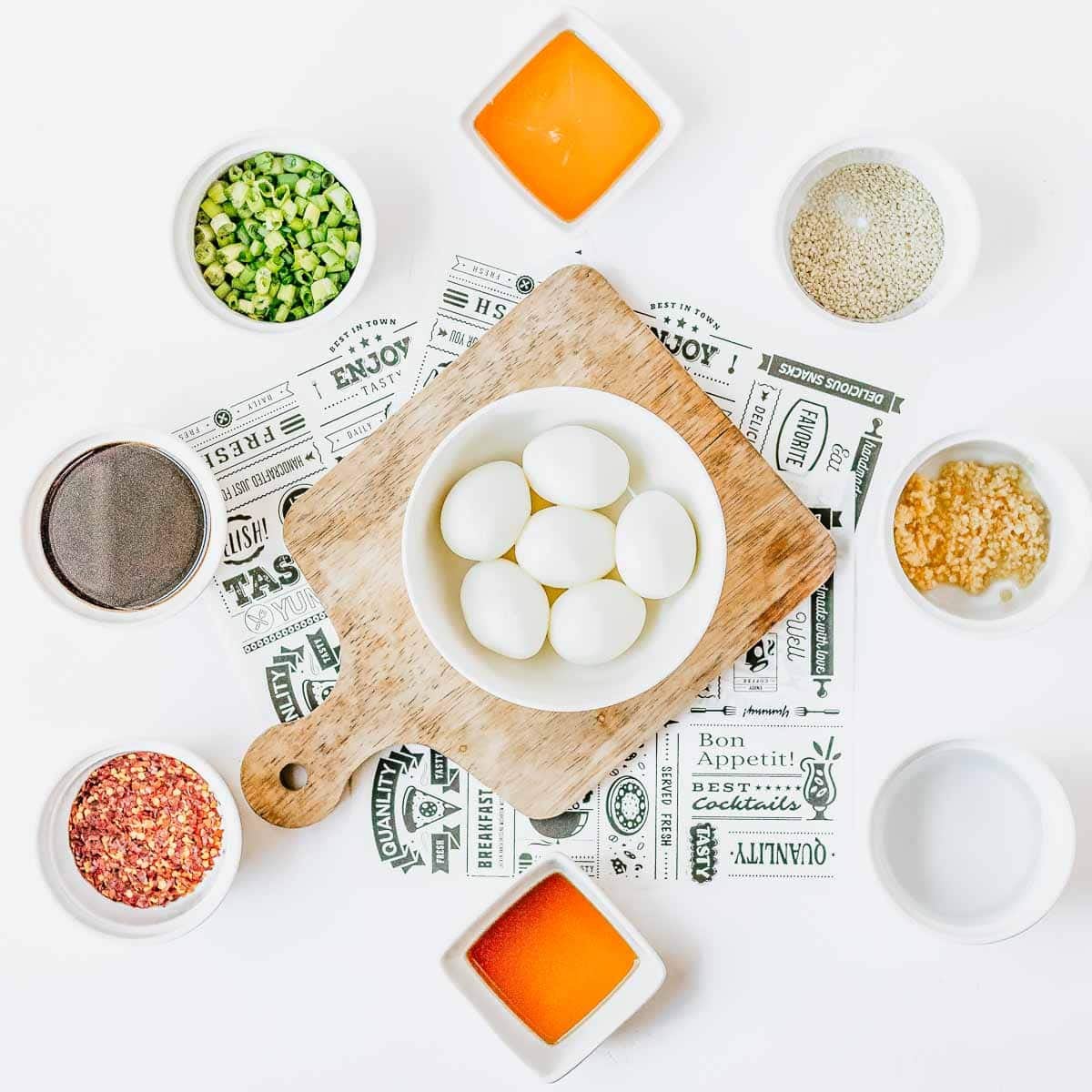 Overhead view of a cutting board with six boiled eggs in a bowl, surrounded by small bowls of chopped green onions, sesame seeds, minced garlic, black pepper, red pepper flakes, and two orange sauces on a white background.
