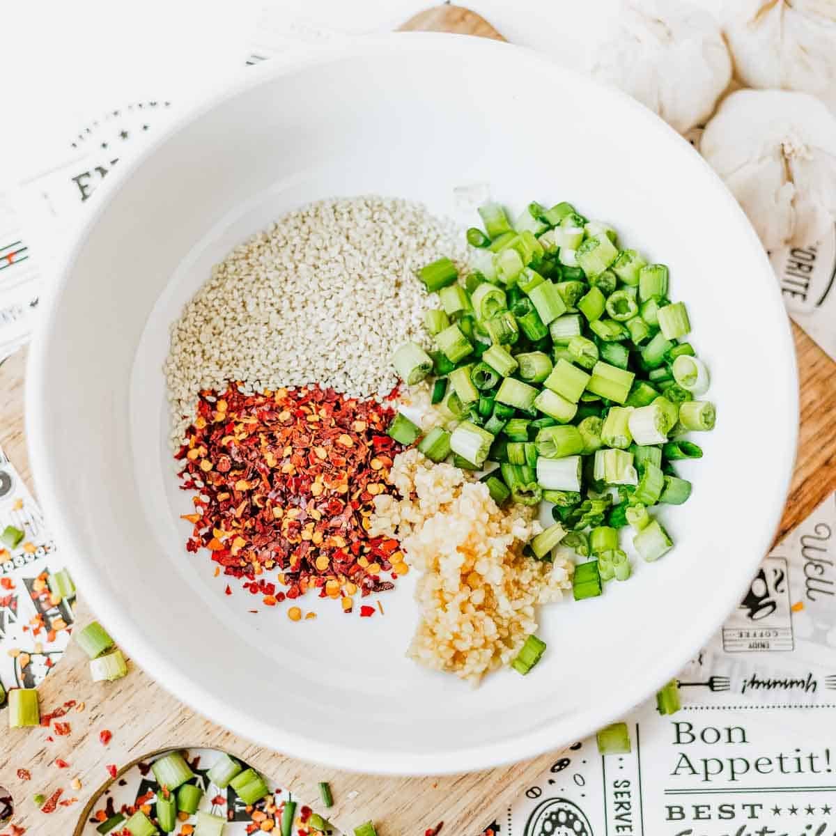 A white bowl contains sesame seeds, red pepper flakes, chopped green onions, and minced garlic, arranged in separate sections. The bowl sits on a wooden surface with scattered green onion slices and a garlic bulb nearby.