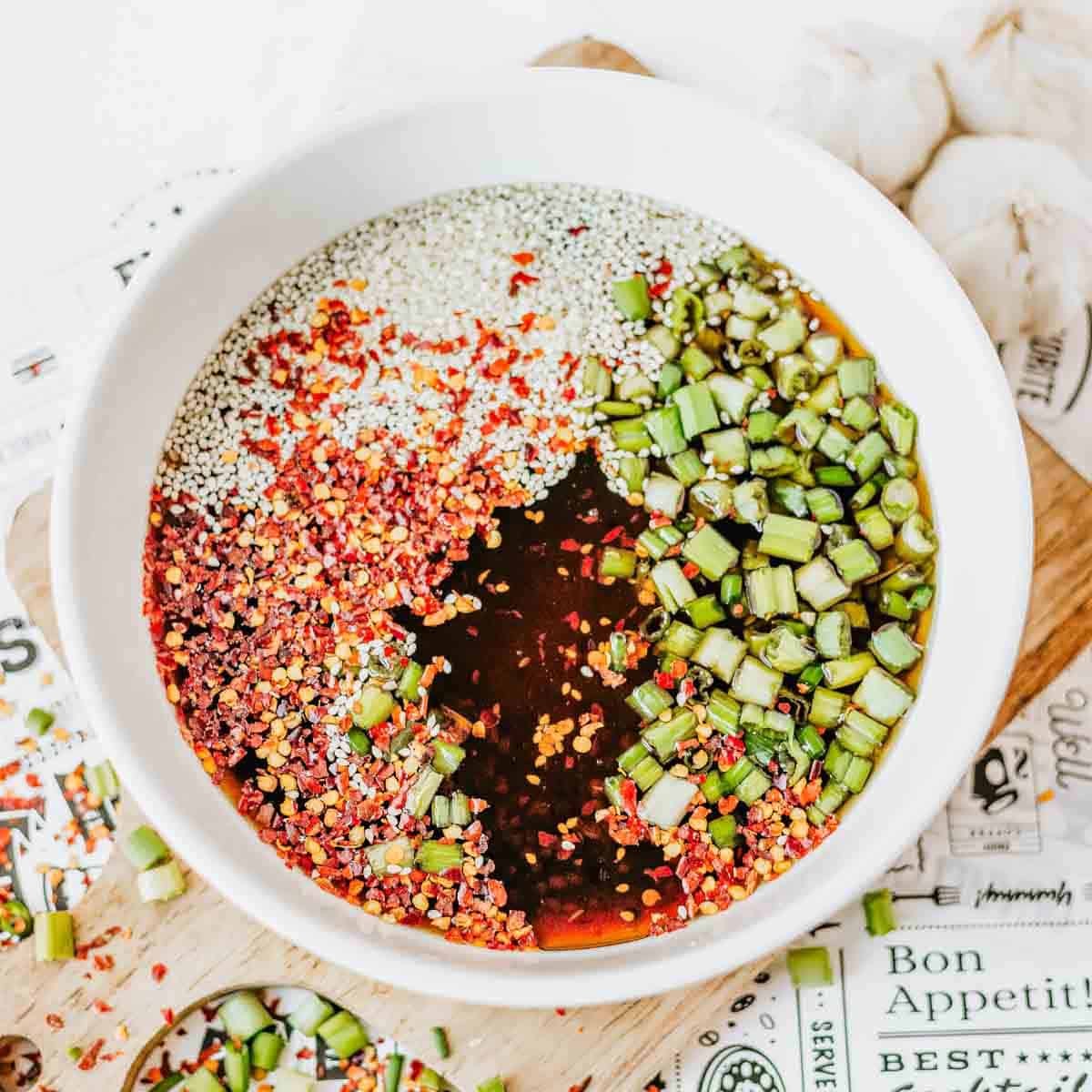 A white bowl filled with a soy sauce mixture containing red chili flakes, sesame seeds, chopped green onions, and a dark liquid, placed on a patterned surface with garlic cloves nearby.