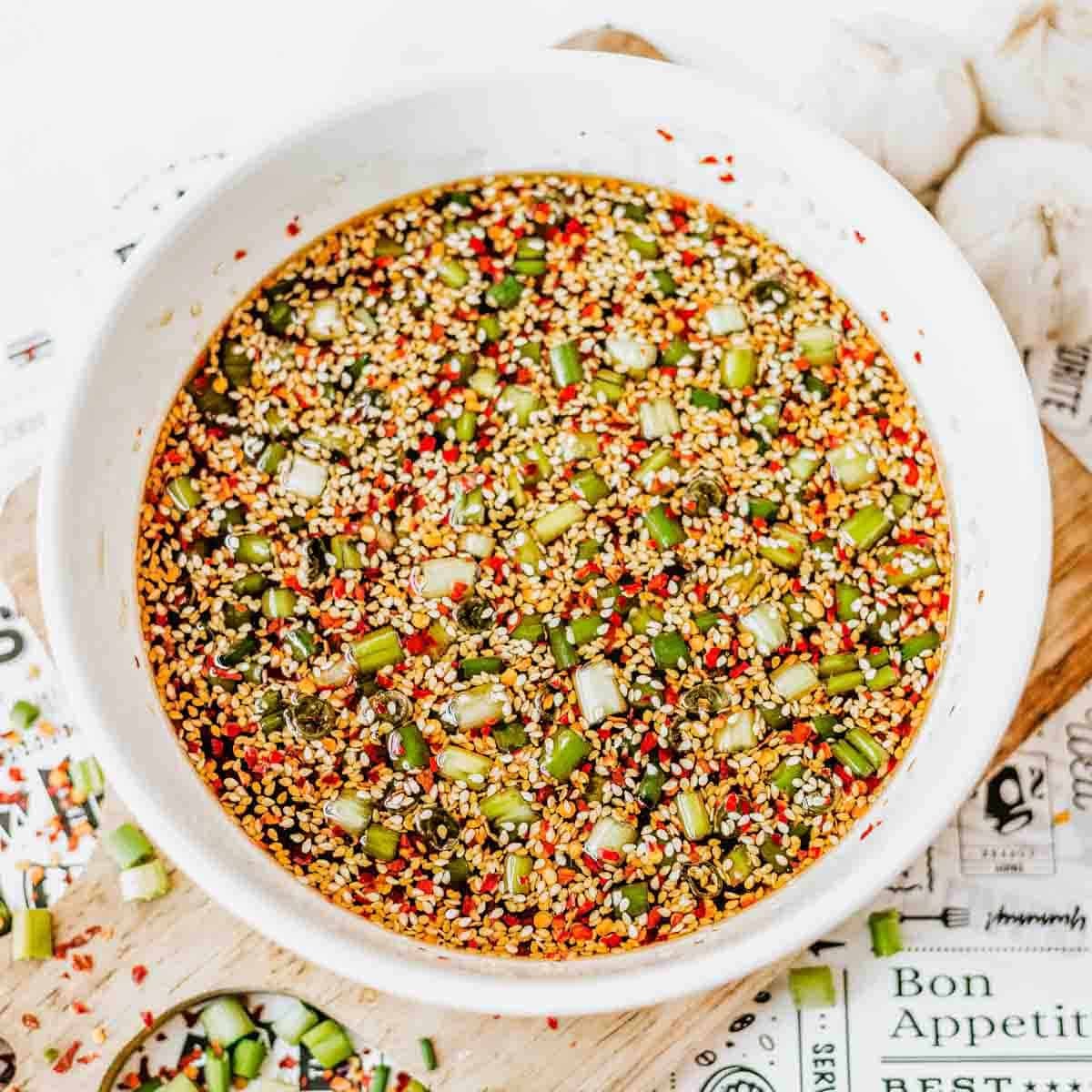 A white bowl filled with a colorful soy sauce marinade containing chopped green onions, sesame seeds, red pepper flakes, and other seasonings. Nearby are pieces of garlic and a cutting board with scattered onions and seasonings.