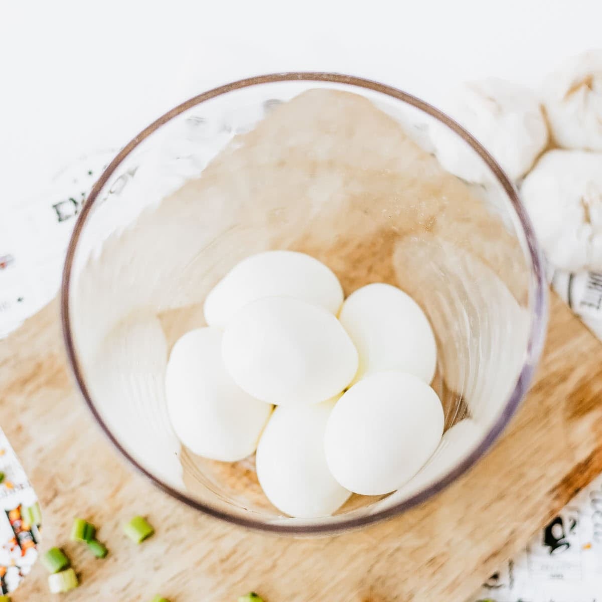 A glass bowl containing five peeled hard-boiled eggs sits on a wooden cutting board next to chopped green onions and garlic cloves.
