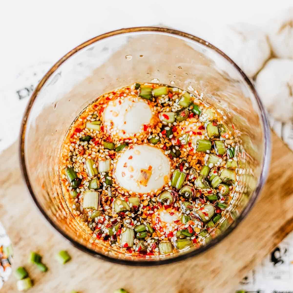 A glass bowl filled with marinated boiled eggs, chopped green onions, sesame seeds, and red chili flakes in a soy-based sauce, placed on a wooden surface.