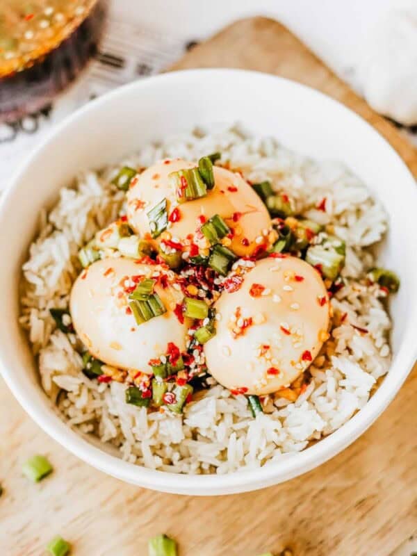A white bowl filled with rice topped with three Korean marinated eggs, garnished with chopped green onions, sesame seeds, and red chili flakes, placed on a wooden board with a fork nearby.